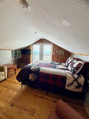 Un dormitorio con piso de madera, una cama con colcha, un aparador de madera, una mesita y una ventana con persianas.