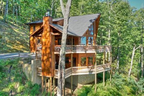 A large, multi-story log cabin with a balcony, surrounded by dense forest.
