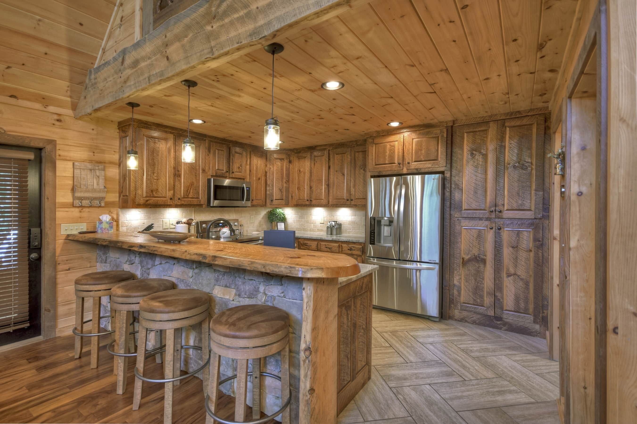 A rustic kitchen with wooden cabinets, a central island with stools, stainless steel appliances, and a herringbone floor pattern.