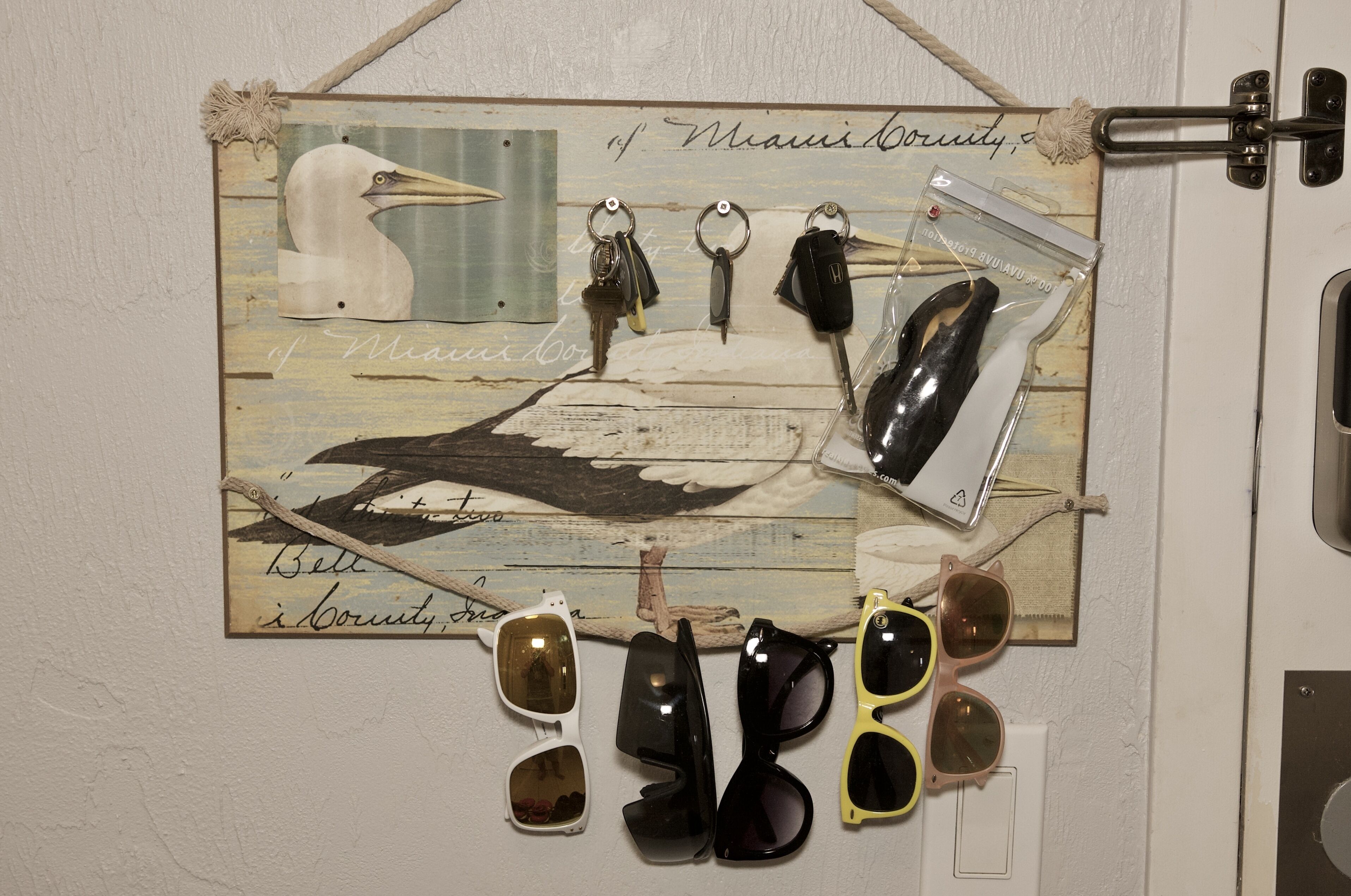 Una pantalla montada en la pared con llaves de estilo antiguo, gafas de sol y una postal que muestra un pájaro y texto escrito a mano.