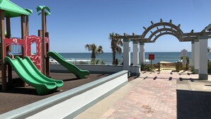 Un parque infantil con un tobogán verde y una estructura de juegos, un camino pavimentado y vistas al océano.
