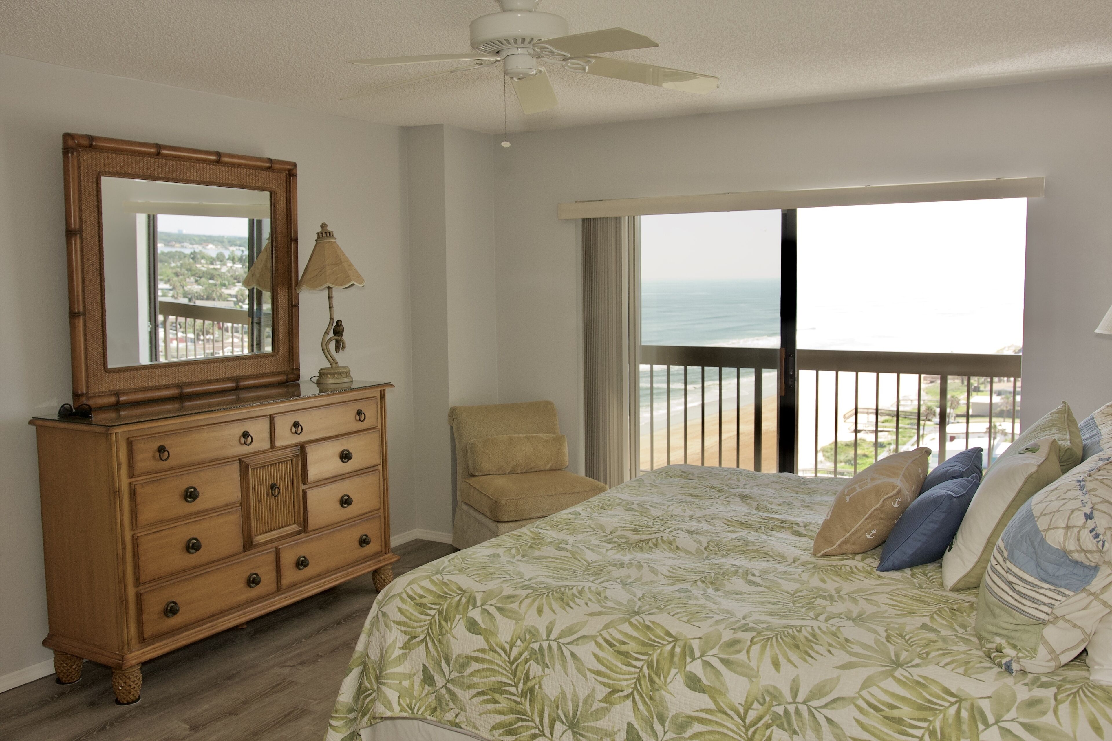 Ein Schlafzimmer mit einem großen Bett, einem Schminktisch, einem Sessel, einem Deckenventilator und einem Balkon mit Meerblick.