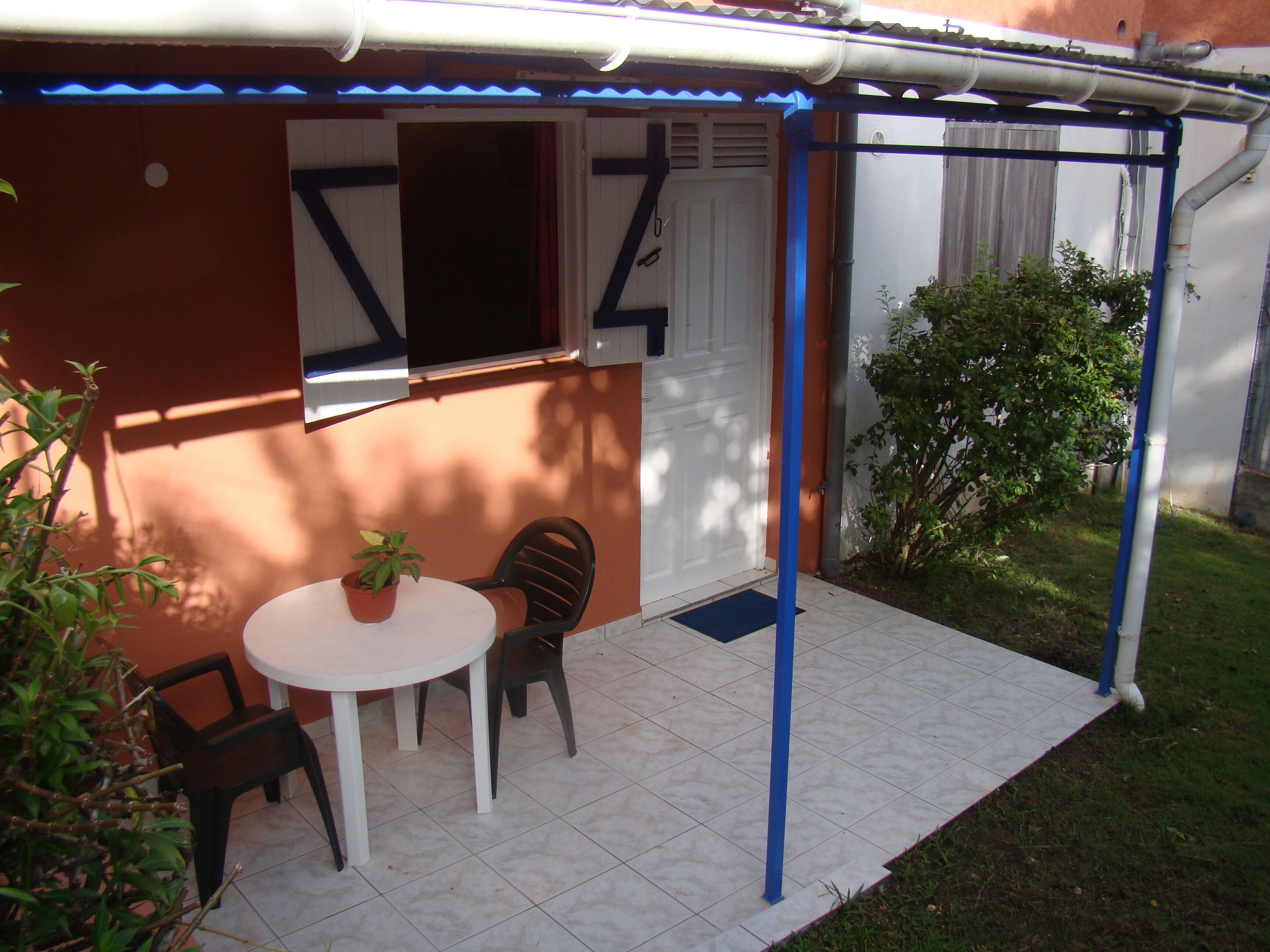 Un piccolo patio con tavolo e sedie, una pianta in vaso e un'area riparata con una porta e una finestra.