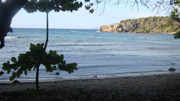 Uma praia com águas cristalinas, um rochedo e vegetação rala.