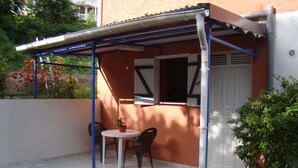 Uma varanda coberta com mesa e cadeiras, um vaso de planta e uma porta com persianas.