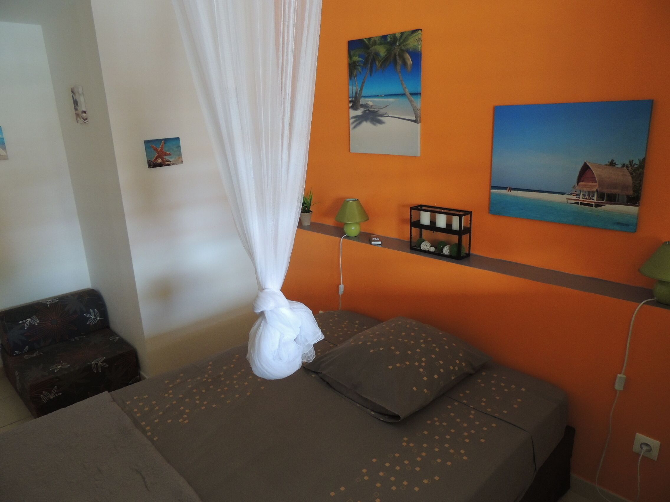 Une chambre avec un mur orange, un lit recouvert d’une couverture grise, un baldaquin blanc, une étagère ornée d’objets décoratifs et un tableau représentant une scène de plage.