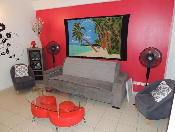 Uma sala de estar com uma parede vermelha, um sofá cinza, duas poltronas, uma mesa de centro de vidro e duas ventoinhas.