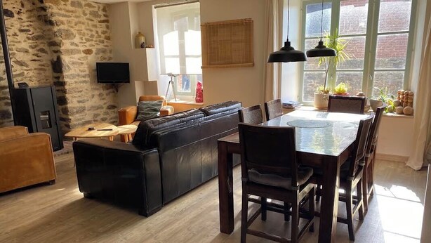 Ein Wohnzimmer mit Steingemäuer, einer schwarzen Ledersofa, einem Holzespeisesaal mit Stühlen und einem Fernseher an der Wand.