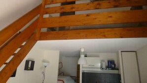 Una habitación con escalera de madera, pared blanca, una mesita con lámpara y una pequeña cocina.