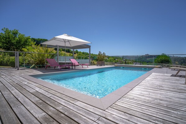 A rectangular swimming pool with a wooden deck, surrounded by pink lounge chairs and a covered seating area. - Villa Calmont : Oasis of Serenity in Aveyron with Private Pool and Jacuzzi (Espalion)