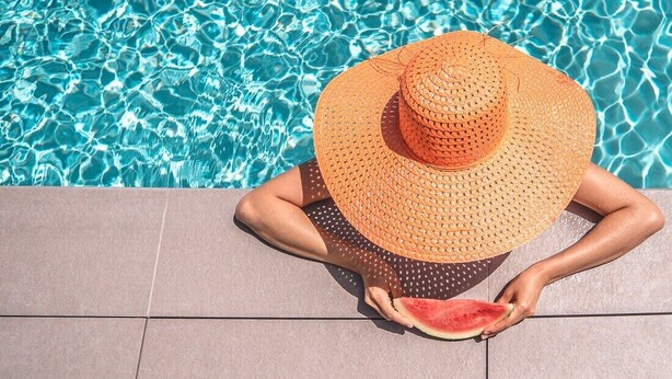Una persona sentada junto a una piscina, con un sombrero de paja grande y sosteniendo una rebanada de sandía.