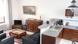 En stue med sofa, sofabord og fjernsyn. Der er et køkkenområde med vask, skabe og komfur. Et billede hænger på væggen, og et vindue med gardiner er synligt.