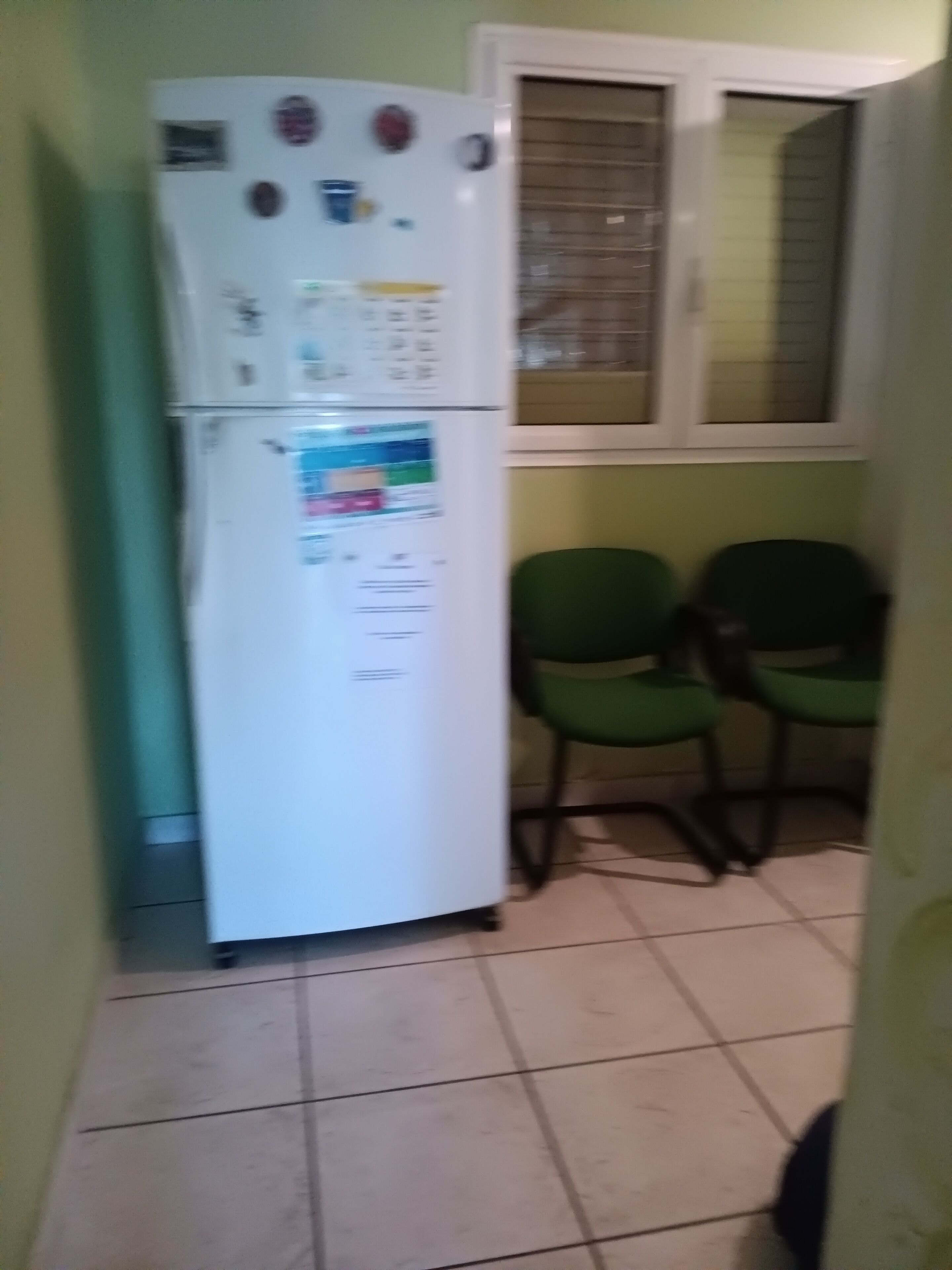 A kitchen with a white refrigerator, two green chairs, and a window with blinds.