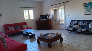 Un salon avec un canapé rouge, un canapé noir orné de motifs végétaux, une table basse en bois, un téléviseur posé sur un meuble et un ventilateur de plafond.