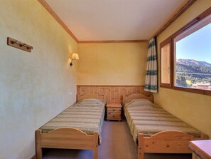 A small room with two wooden beds, a nightstand, a wall-mounted lamp, and a window with a view of mountains.