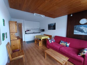 A compact living space with a kitchenette, a red sofa with patterned cushions, and a wooden dining table. - Apartment for 4, near slopes and shops, parking (Montgenèvre)
