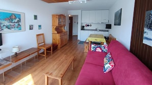 Uma sala de estar acolhedora com teto de madeira, um sofá vermelho, uma mesa de jantar de madeira e uma cozinha ao fundo.