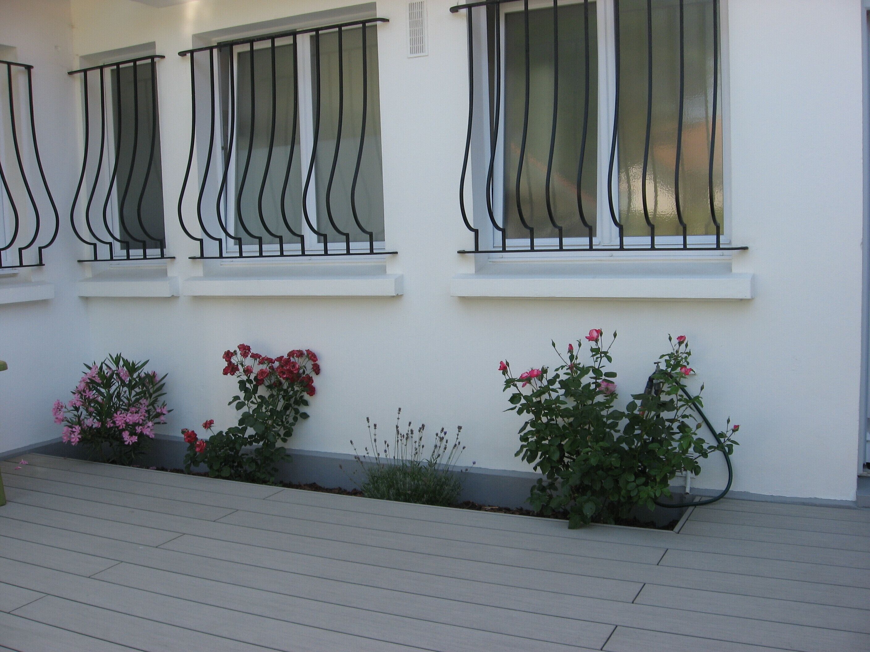 A building facade with three windows featuring black metal railings and white frames. There are potted plants with pink flowers and green foliage along the base of the windows.
