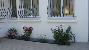 Une façade d’immeuble avec trois fenêtres, dotées de balustrades en métal noir et de cadres blancs. Des plantes en pot, avec des fleurs roses et un feuillage vert, sont disposées le long du bas des fenêtres.