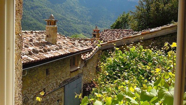 Un bâtiment rustique en pierre, doté d’un toit en tuiles et de cheminées, entouré d’une végétation luxuriante et avec des montagnes en arrière-plan.