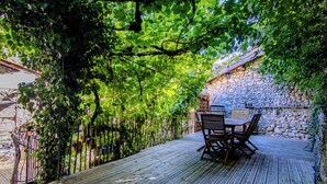 Une terrasse en bois avec une table et des chaises, entourée de verdure et d’un mur en pierre.