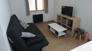 Un salon moderne avec un canapé noir, une table basse blanche, un téléviseur posé sur un meuble en bois et une fenêtre avec des rideaux.