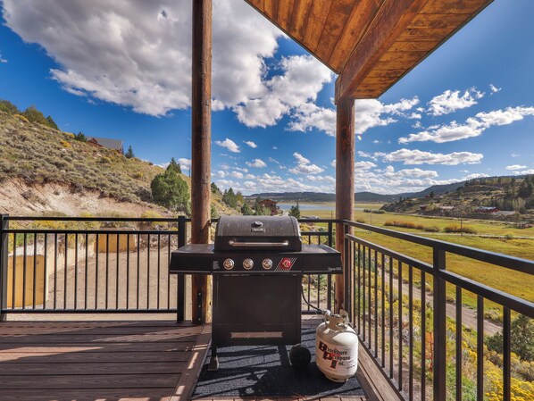 A barbecue grill on a wooden deck with a scenic view of hills and a lake.
