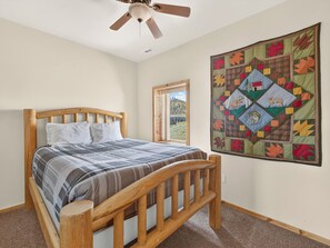 A bedroom with a wooden bed, a quilt, a ceiling fan, a window with a view, and a large wall hanging.