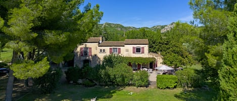 A two-story house with a garden, surrounded by trees and hills in the background.