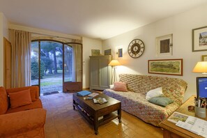 A cozy living room with a sofa, a coffee table, and a lamp. There is a large window with curtains, a clock on the wall, and a door leading outside.