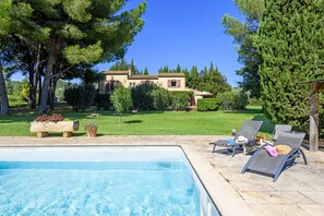 A swimming pool with lounge chairs and a stone bench, surrounded by greenery and a house in the background.