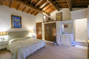 A bedroom with a wooden ceiling, a bed with a floral bedspread, a dresser, a vanity with a mirror, and a shower area.