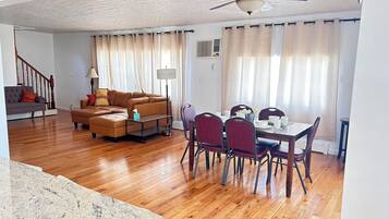 A spacious living area with a wooden floor, a brown sectional sofa, a dining table with red chairs, and a ceiling fan.