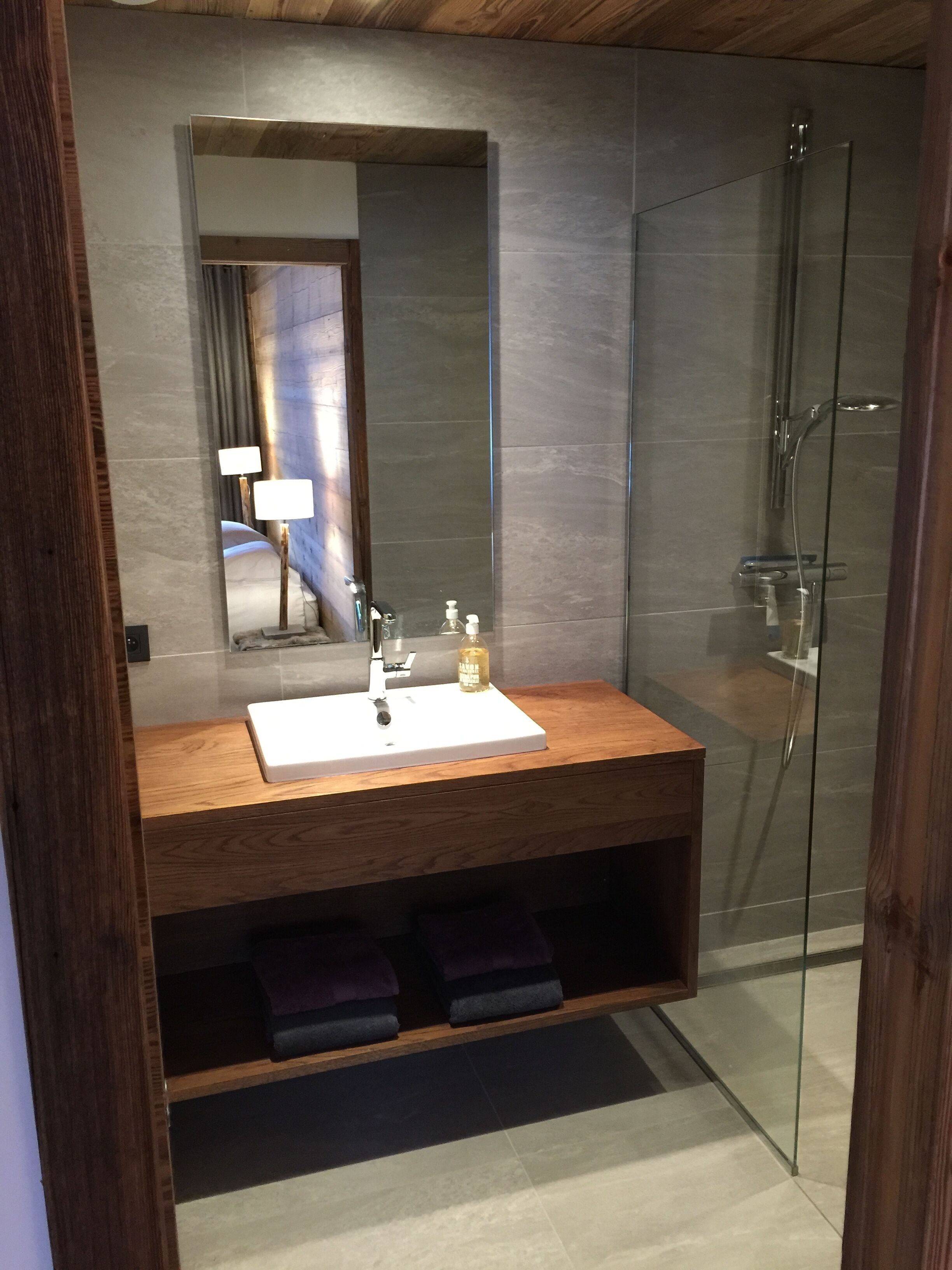 A modern bathroom with a wooden vanity, a large mirror, a glass shower enclosure, and a built-in shelf with towels.