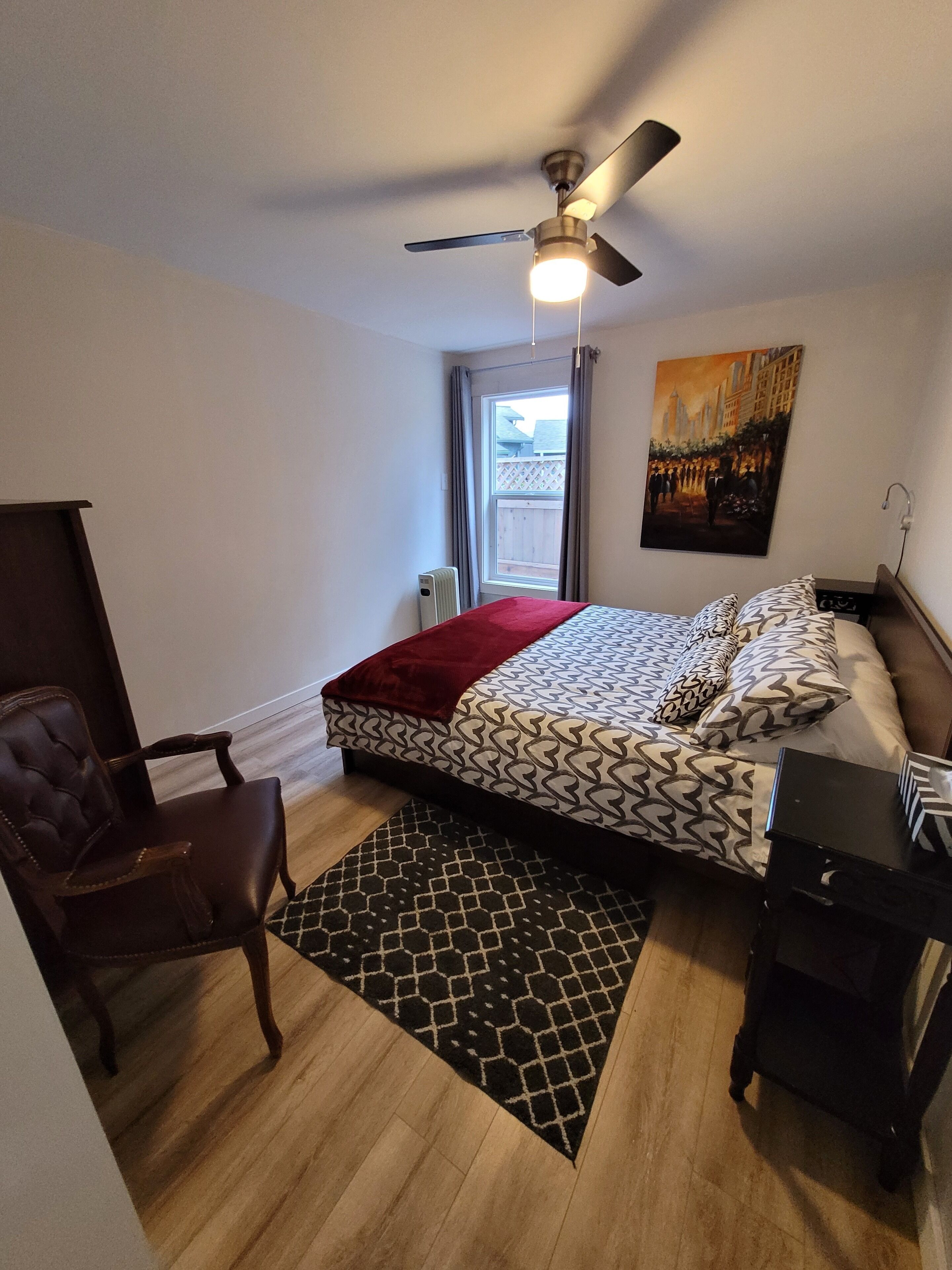Une chambre à coucher comprenant un lit, une chaise, un bureau, un ventilateur de plafond, un radiateur, une fenêtre et un tableau.