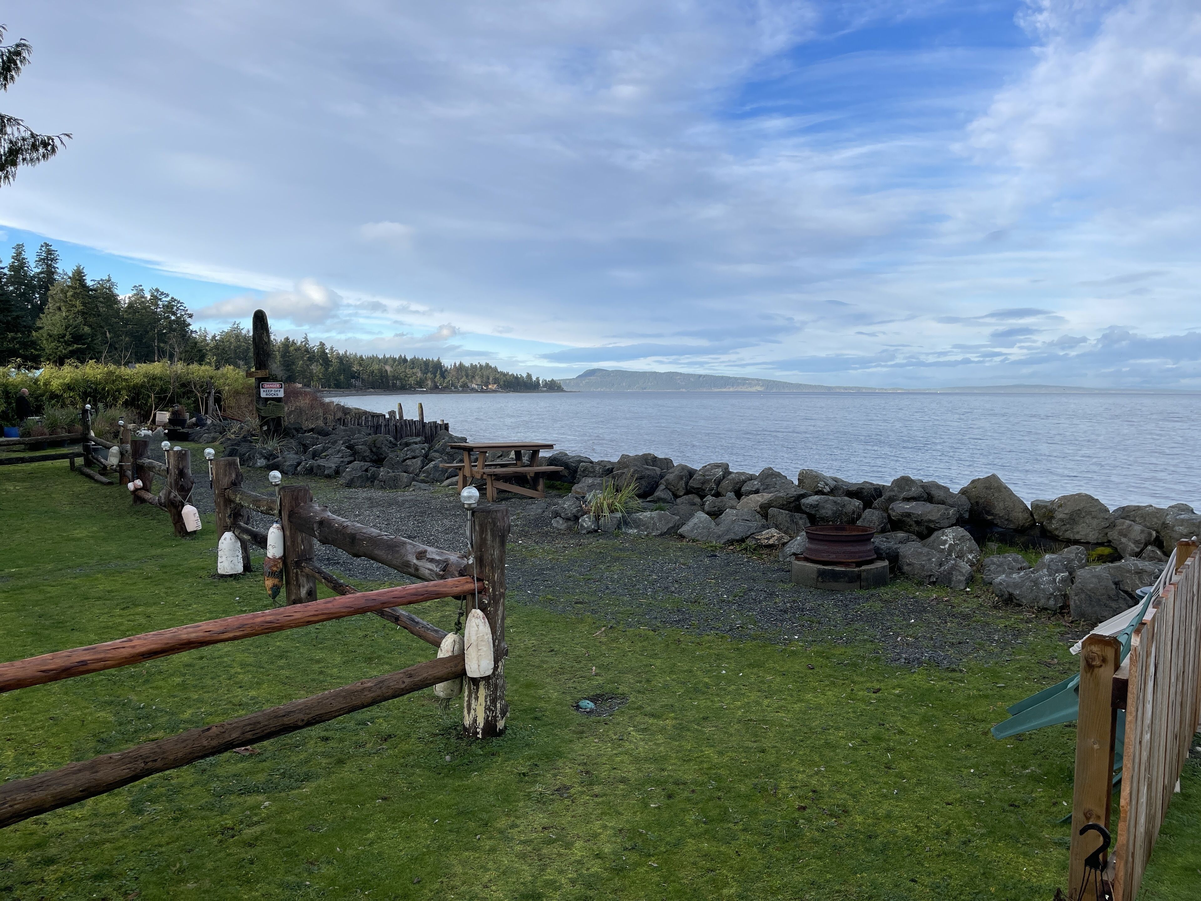 A waterfront park with picnic tables, a wooden fence, and a rocky shoreline.