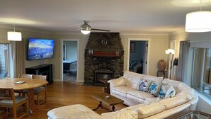 Una sala de estar con chimenea de piedra, un televisor, un comedor con mesa y sillas de madera, y un ventanal con cortinas.