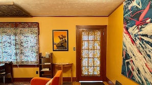 Een kamer met een houten vloer, oranje stoelen, een bureau met stoel, een boekenkast, een ingelijst poster, een deur met gordijn met patroon, en een groot abstract schilderij aan de muur.