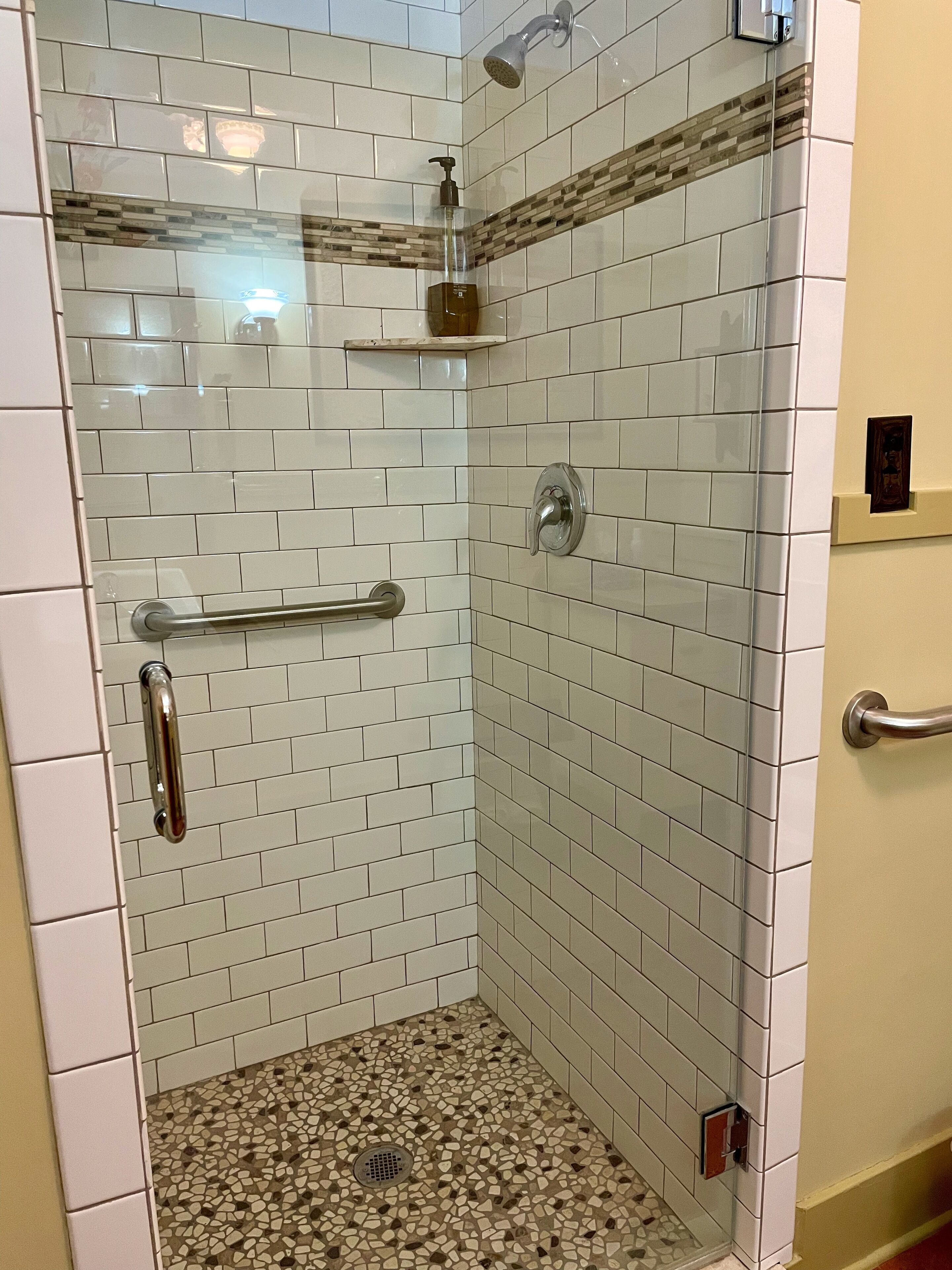 Eine Dusche mit weißen Fliesen, einer Glastür und einer Haltestange.
