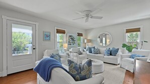 Un salón amplio con muebles blancos, ventilador de techo y ventanas grandes con persianas.