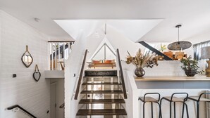 Un interior moderno con una escalera de madera, paredes blancas y una zona de cocina con taburetes de bar.