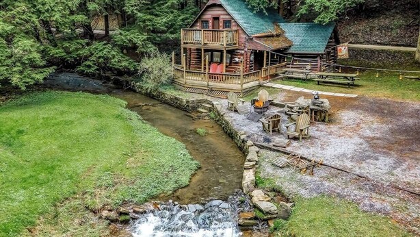 Une cabane dotée d’un porche et d’une petite terrasse, entourée d’une forêt et bordée par un ruisseau.