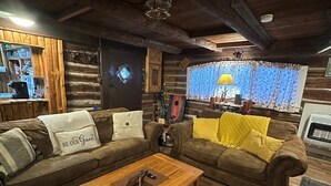 L’intérieur d’un chalet confortable, avec un plancher en bois, deux canapés marron ornés de coussins, une table basse en bois et une décoration rustique comprenant une lampe et une fenêtre avec des rideaux.