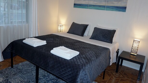 Een keurig opgemaakt bed met een donkere gewatteerde spreidek en twee kussens, een nachtkastje met een lamp erop, en een raam met dunne gordijnen.