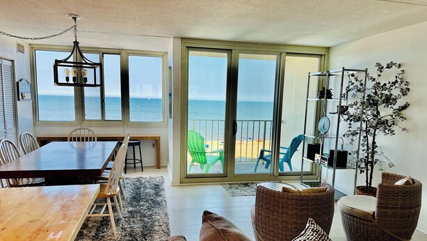 Ein geräumiges Wohnzimmer mit Esstisch aus Holz, bequemen Sitzgelegenheiten, einem großen Fenster mit Blick auf den Strand und einem Balkon mit Meerblick.
