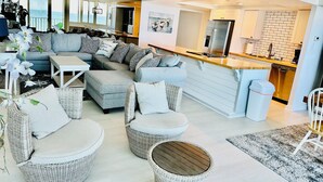 Ein modernes Wohnzimmer mit einem grauen Ecksofa, Korbstühlen und einem Essbereich, im Hintergrund ist eine Küche zu sehen.