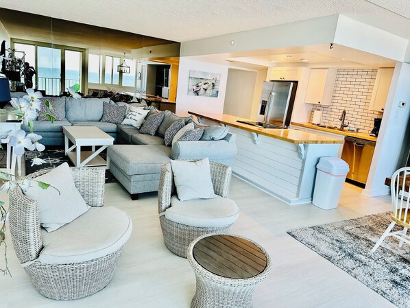 A modern living room with a grey sectional sofa, wicker chairs, and a dining area with a kitchen in the background.