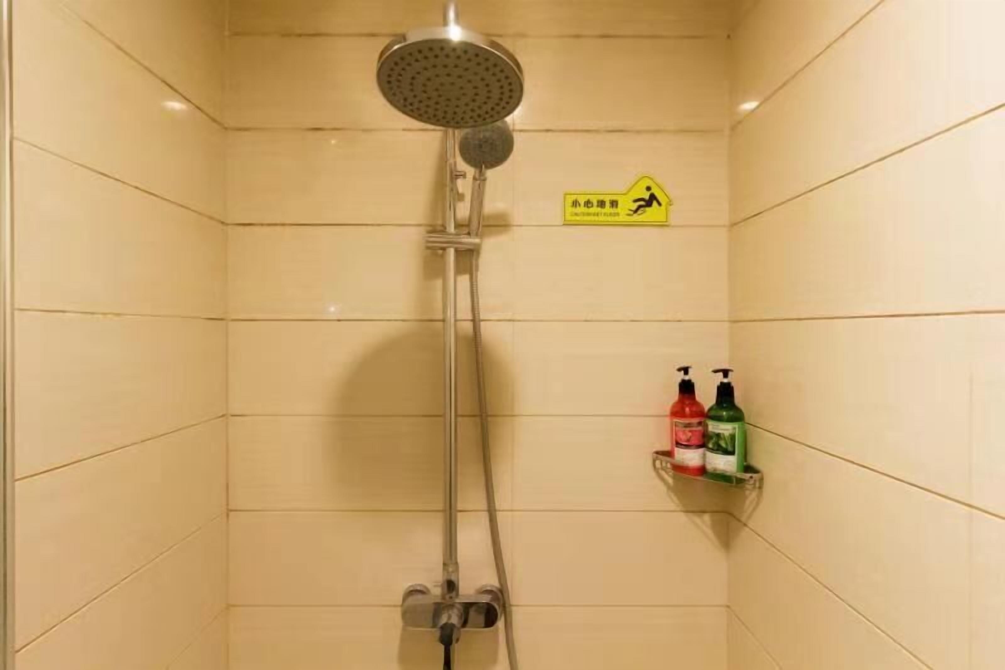 Ducha con cabezal de ducha de mano y grifo de pared. Hay un letrero amarillo con un símbolo de peatón y texto. Dos botellas de champú y acondicionador están en un estante.
