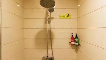 Eine Dusche mit Handbrause und Armaturen an der Wand. Ein gelbes Schild mit Piktogramm für Fußgänger und Beschriftung. Auf einem Regal stehen zwei Flaschen Shampoo und Conditioner.