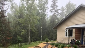 Un patio con mobiliario exterior y una chimenea, rodeado de un paisaje boscoso y una casa al fondo.